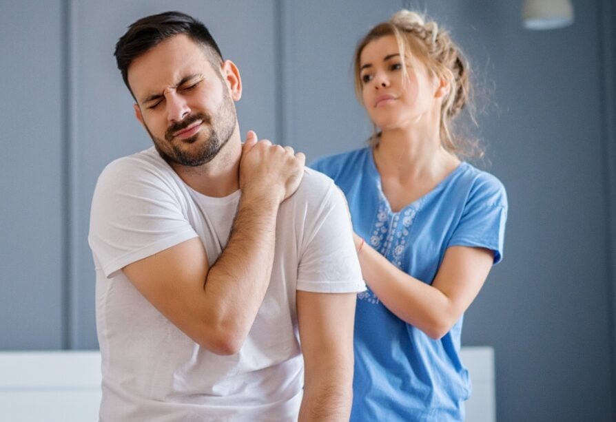 Man sitting up in bed clutching his left trapezius area while his wife tries to help with some massage
